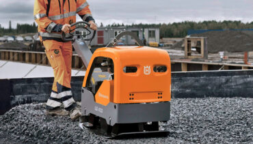 A construction worker pushing an orange Husqvarna plate compactor across gravel on a building site.