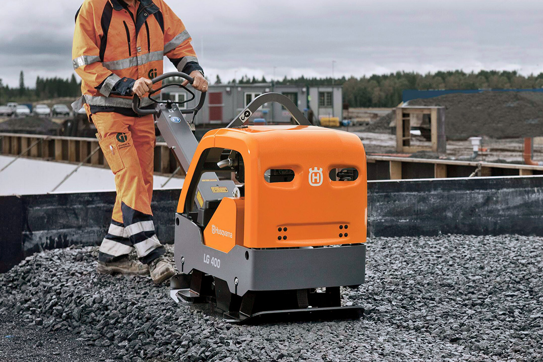 A construction worker pushing an orange Husqvarna plate compactor across gravel on a building site.