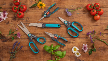 The complete range of GARDENA scissors and snips on a chopping board surrounded by herbs and flowers.