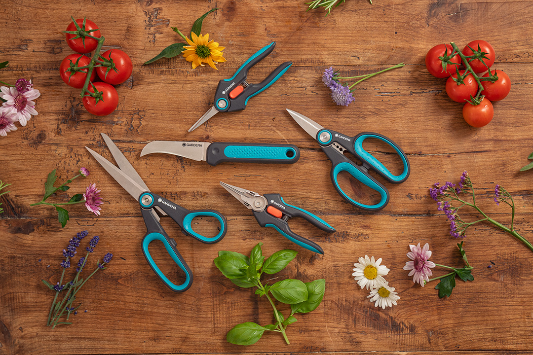 The complete range of GARDENA scissors and snips on a chopping board surrounded by herbs and flowers.