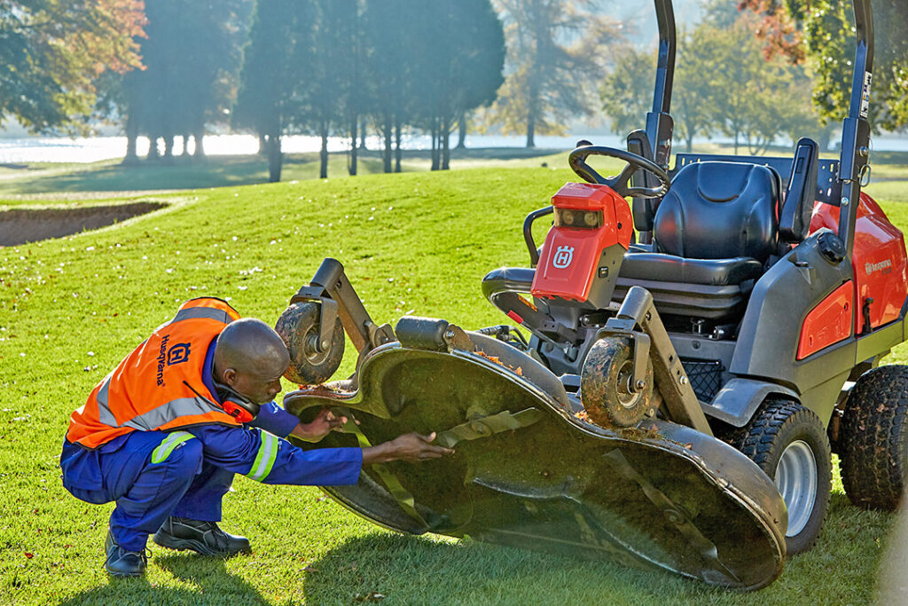 A man checks the blades and cutting deck on a Husqvarna ride-on mower.