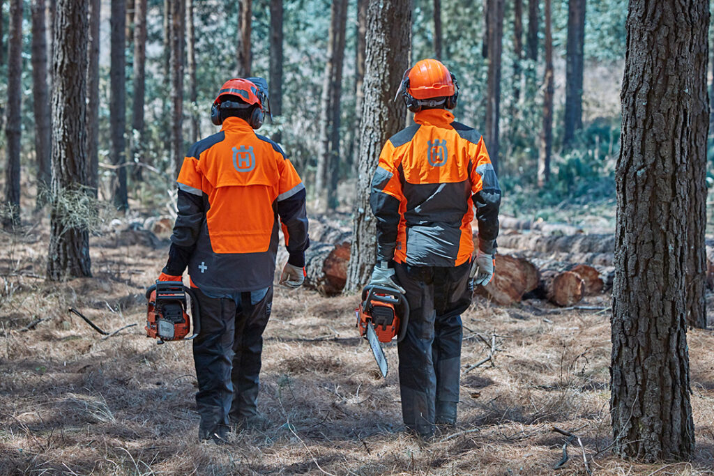 Two treefellers wearing Husqvarna high-vis jackets and forestry helmets carrying Husqvarna chainsaws.