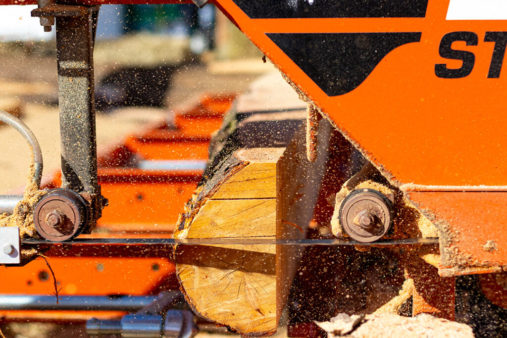 The blade of a Wood-Mizer sawmill cutting through a large log while sawdust floats in the air.